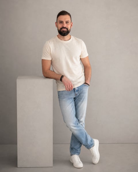 Bearded man in white t-shirt and blue jeans leaning against white block on neutral background